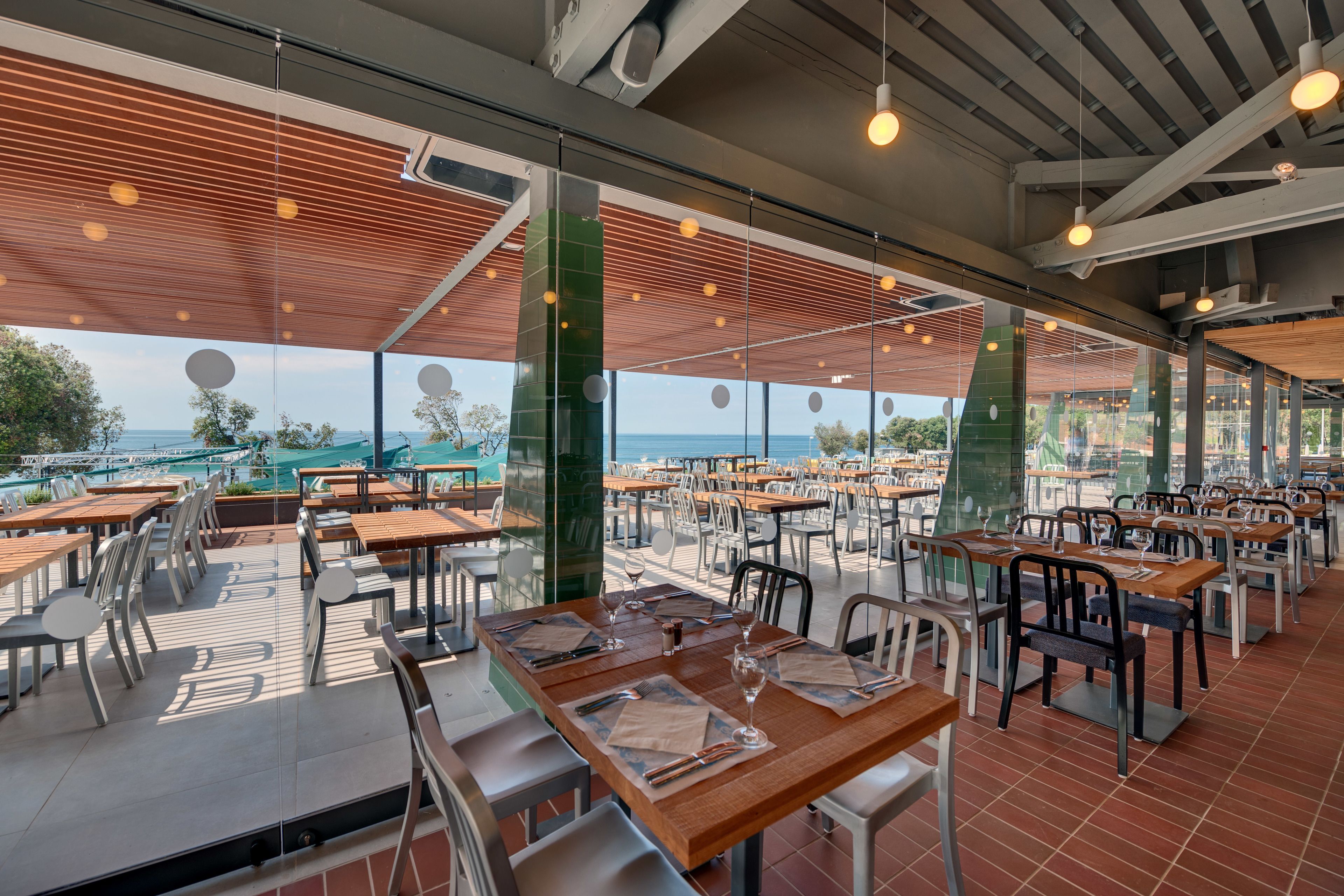 Un vaste espace extérieur aménagé pour les repas, avec de nombreuses tables et chaises, de grandes fenêtres offrant une vue sur la mer, et un design architectural moderne avec des poutres en bois et des luminaires suspendus.