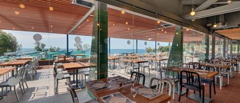 Un amplio comedor al aire libre con varias mesas y sillas, grandes ventanales que ofrecen vistas al mar, y un diseño arquitectónico moderno con vigas de madera y luces colgantes.