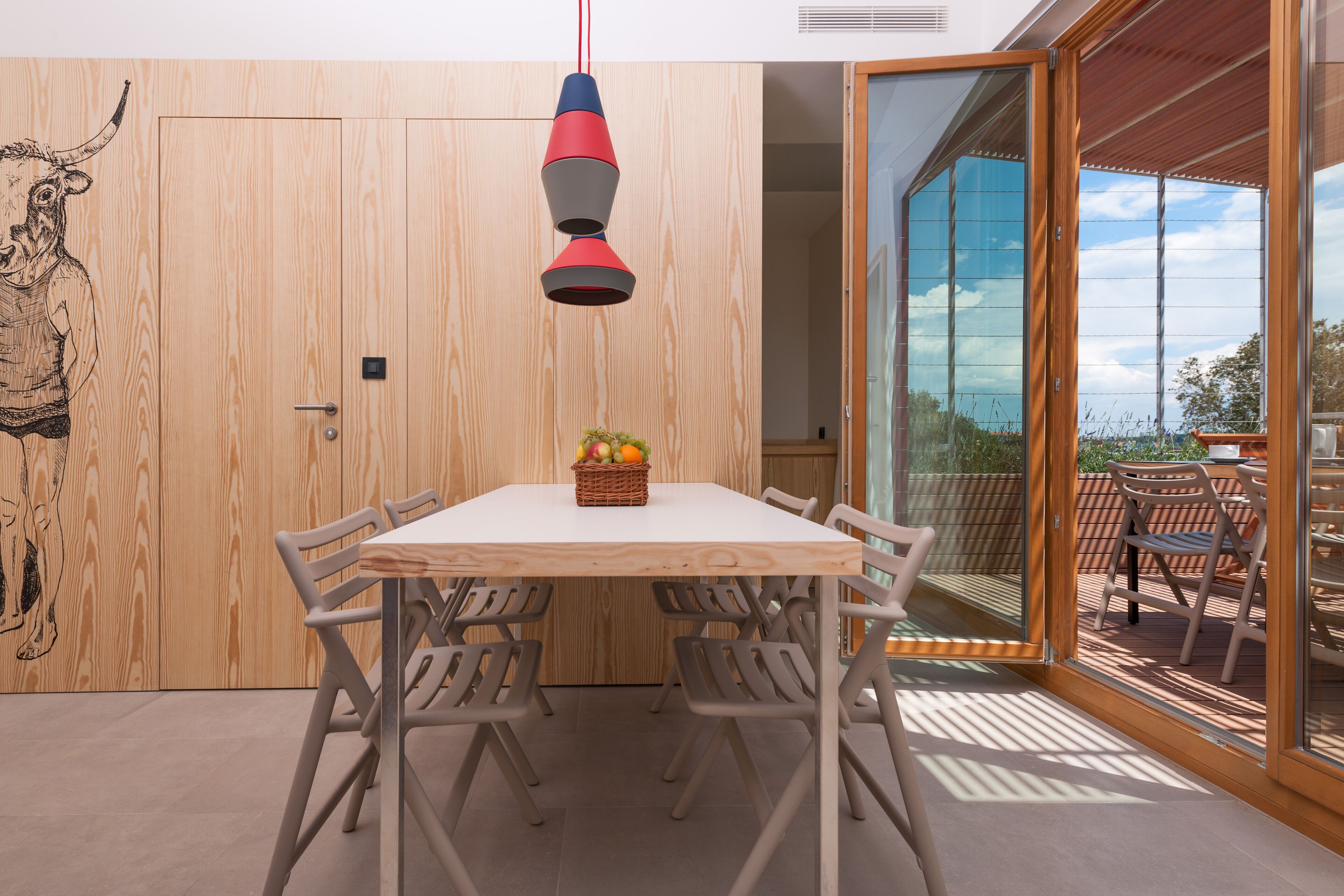 A dining area with a wooden table and chairs, a hanging light fixture, and a view of the outdoors.