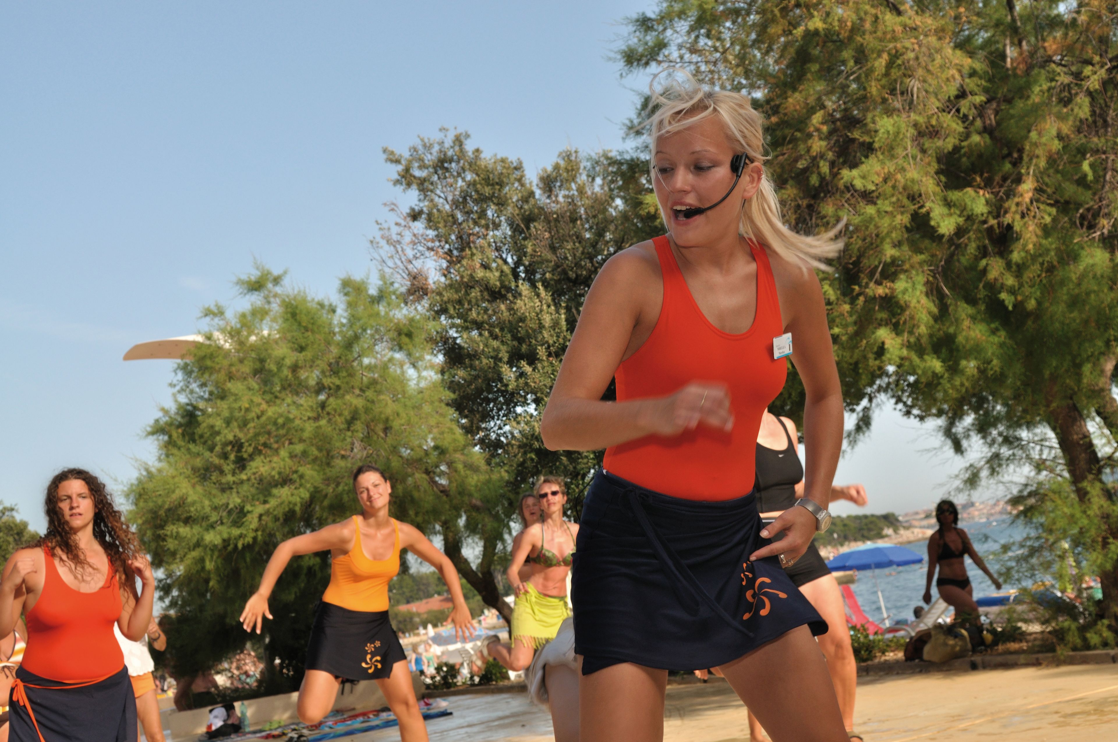 En gruppe mennesker træner udendørs, sandsynligvis i en park eller rekreativt område med træer og et vandareal i baggrunden.