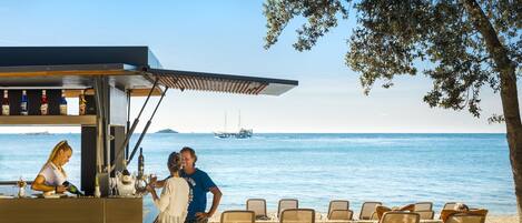 A beachside bar with a view of the sea, outdoor seating, and a bartender.