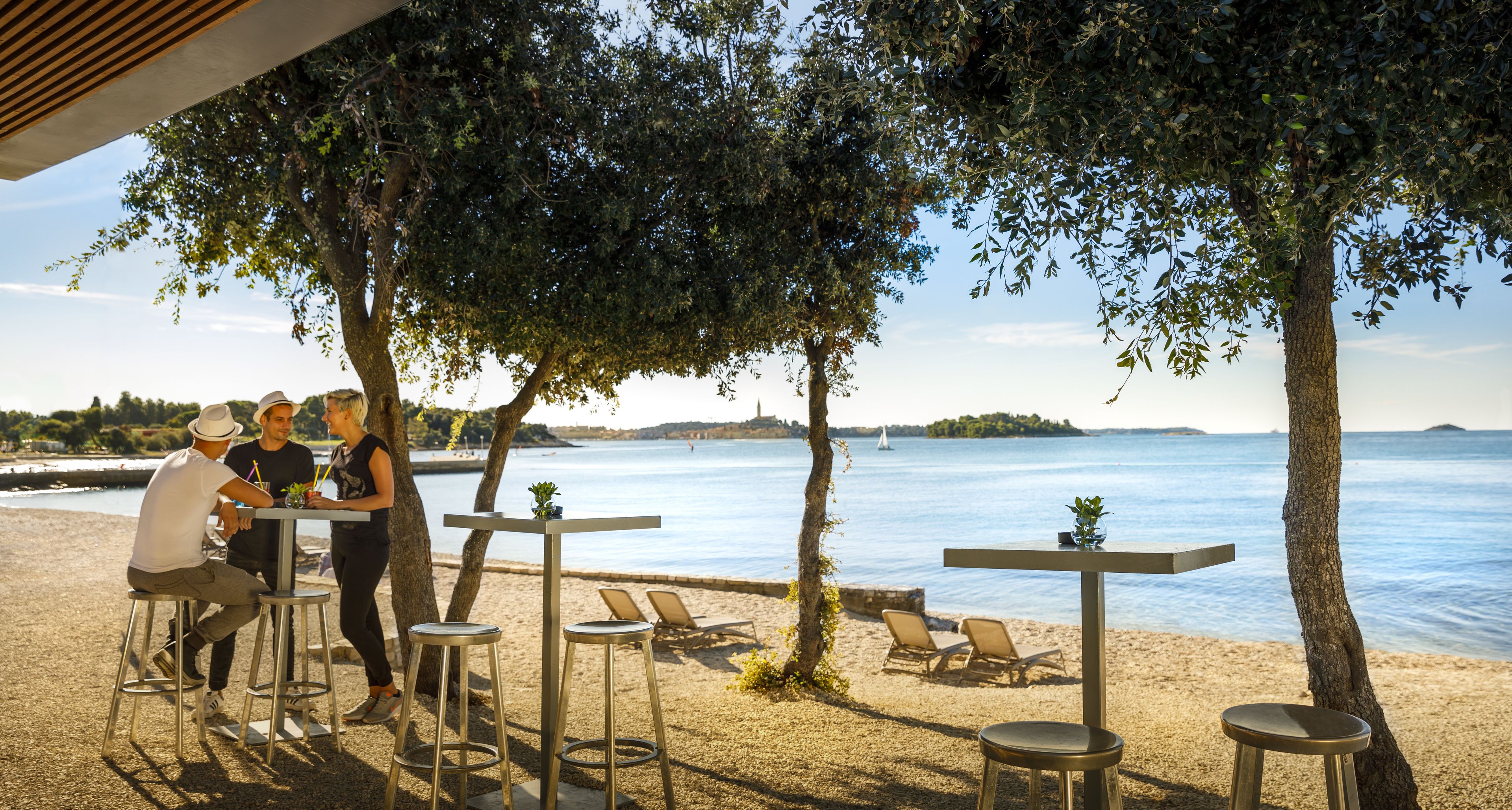 Un área de descanso frente a la playa con mesas y sillas bajo los árboles, con vistas a un mar tranquilo.