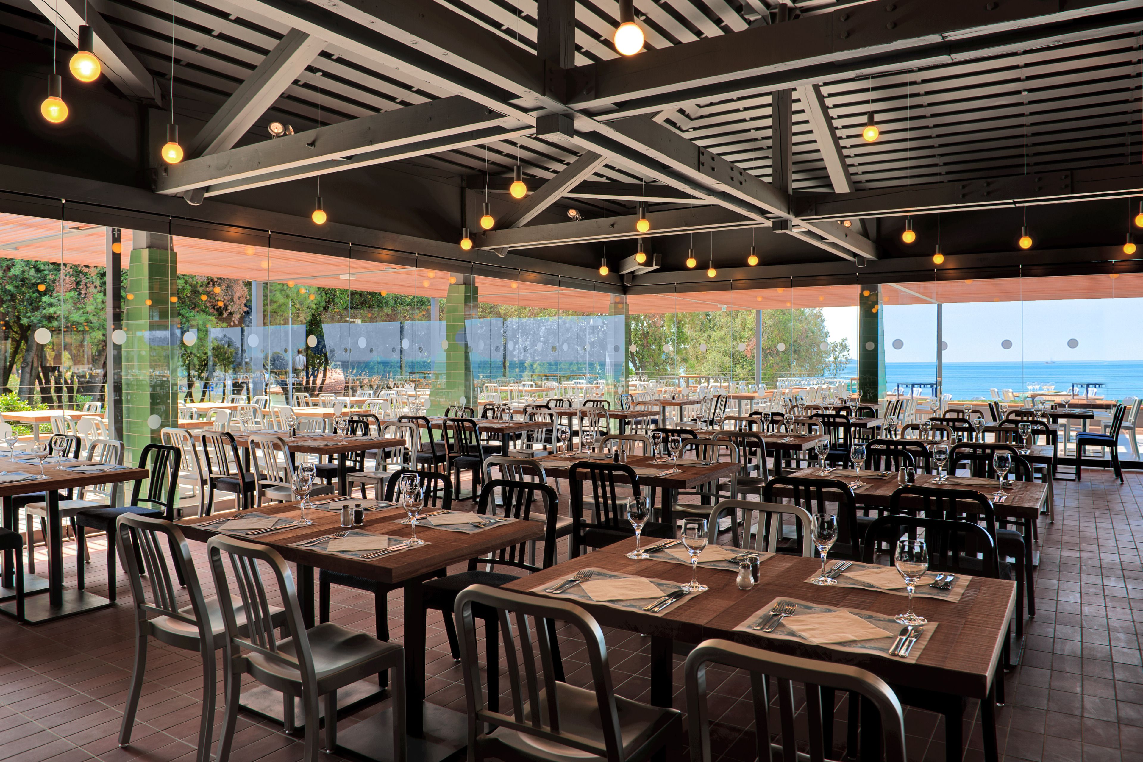 Un ristorante con tavoli all'aperto, apparecchiati con posateria e bicchieri, e vista sul mare.