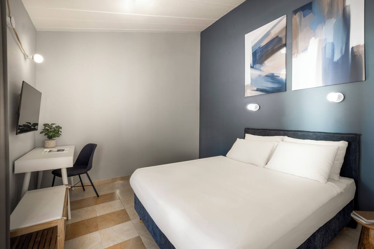 Quarto de hotel moderno com uma cama grande, uma escrivaninha com cadeira, uma mesa pequena e uma planta em vaso.