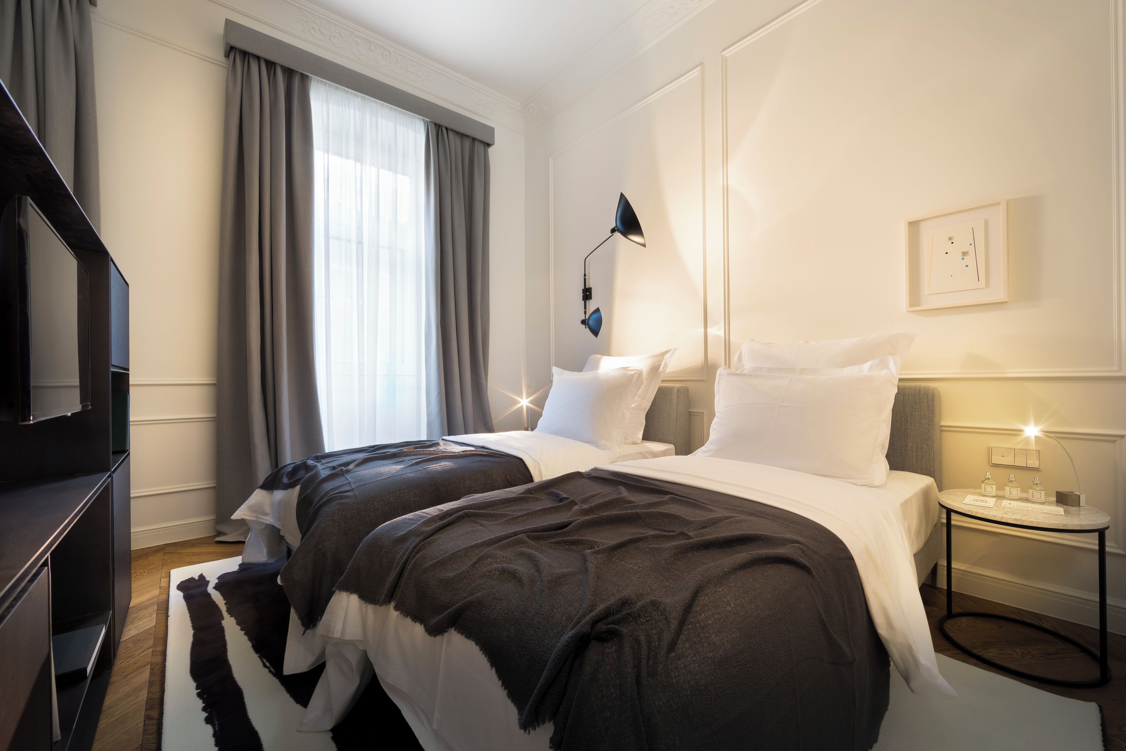 Moderni hotellihuone, jossa on kaksi sänkyä, televisio, yöpöytä lampun kanssa ja ikkuna, jossa on verhot.