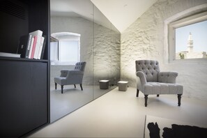 A modern room with a large mirror, two grey armchairs, a bookshelf, and a window with a view of a tower.