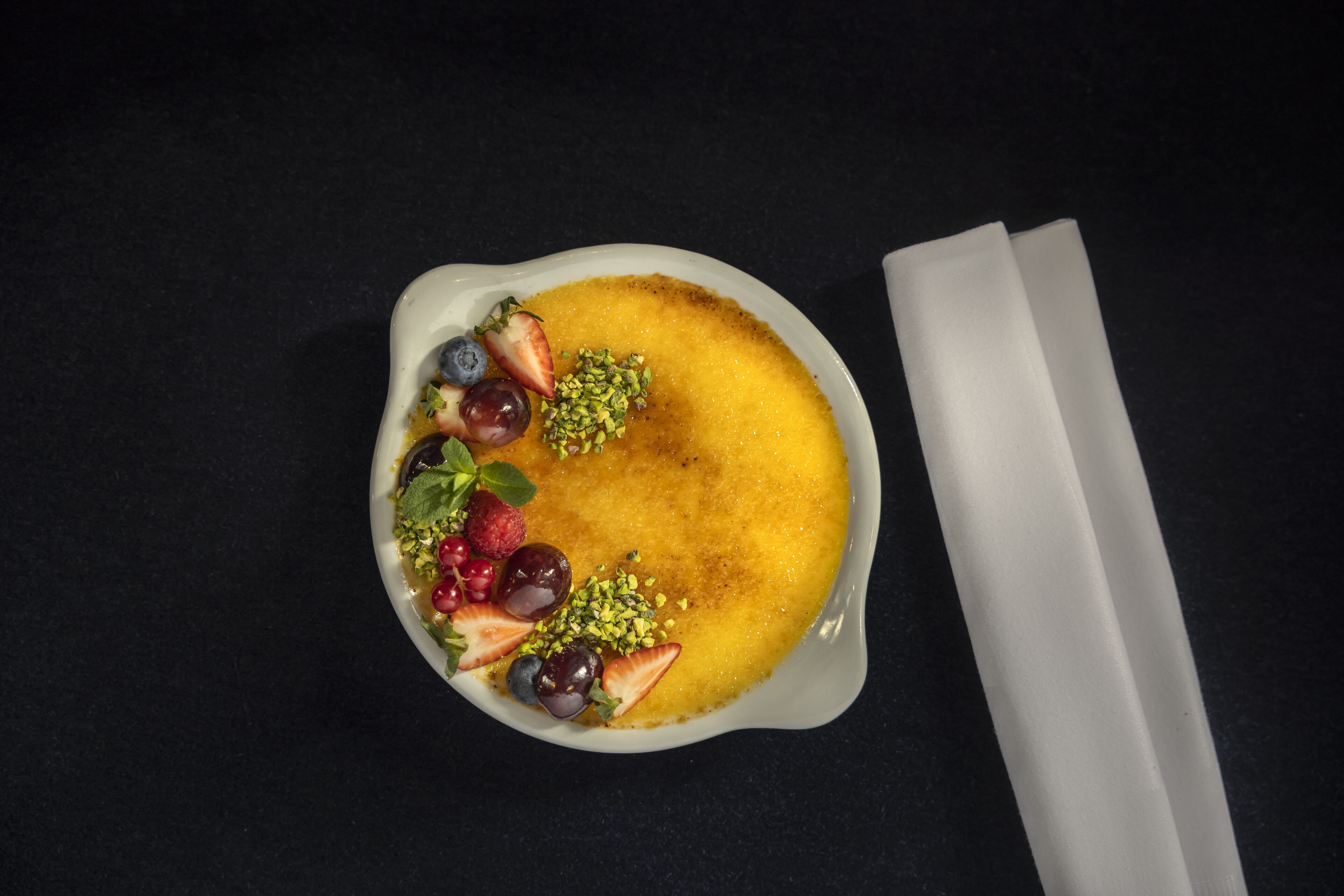 A dessert in a white bowl with strawberries, grapes, and pistachios on a dark surface.