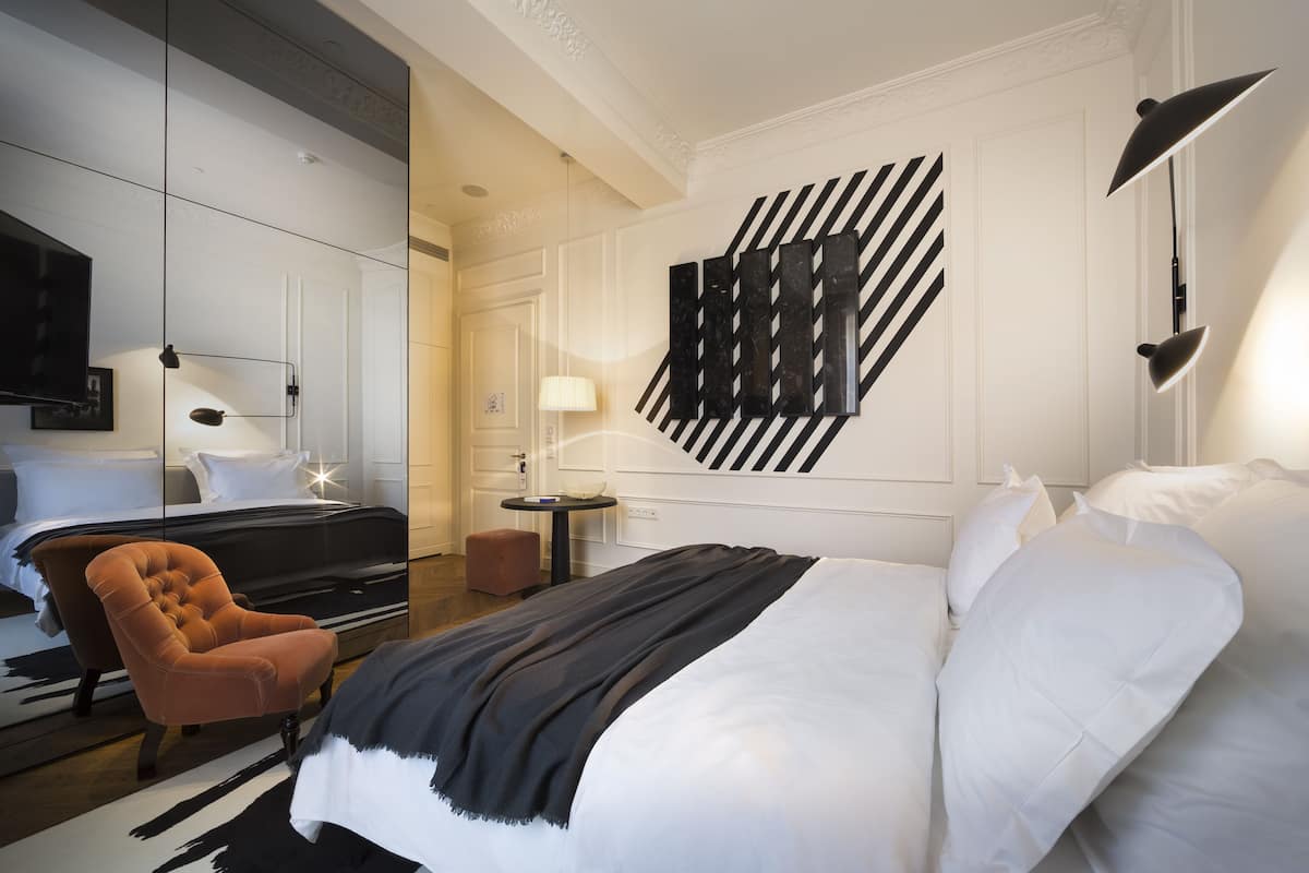A modern hotel room with a large bed, a tufted armchair, a round side table, and a wall-mounted TV.
