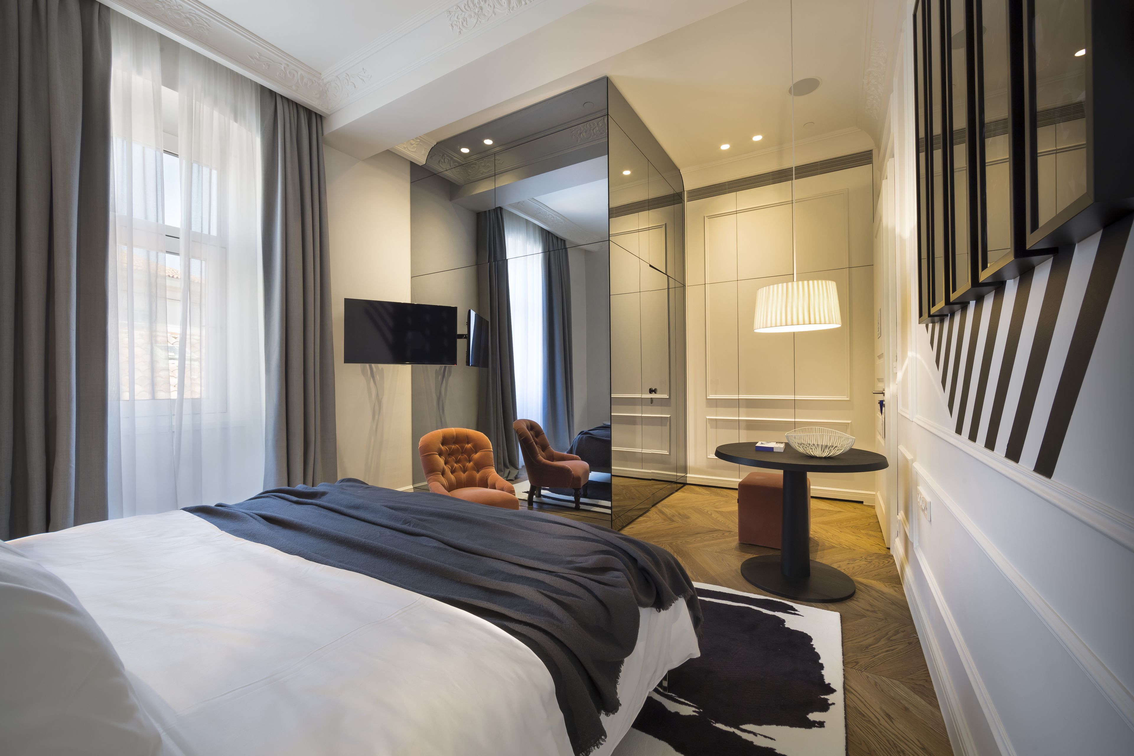A modern hotel room with a large bed, a flat-screen TV, a seating area with two chairs, a small table, and a mirror.