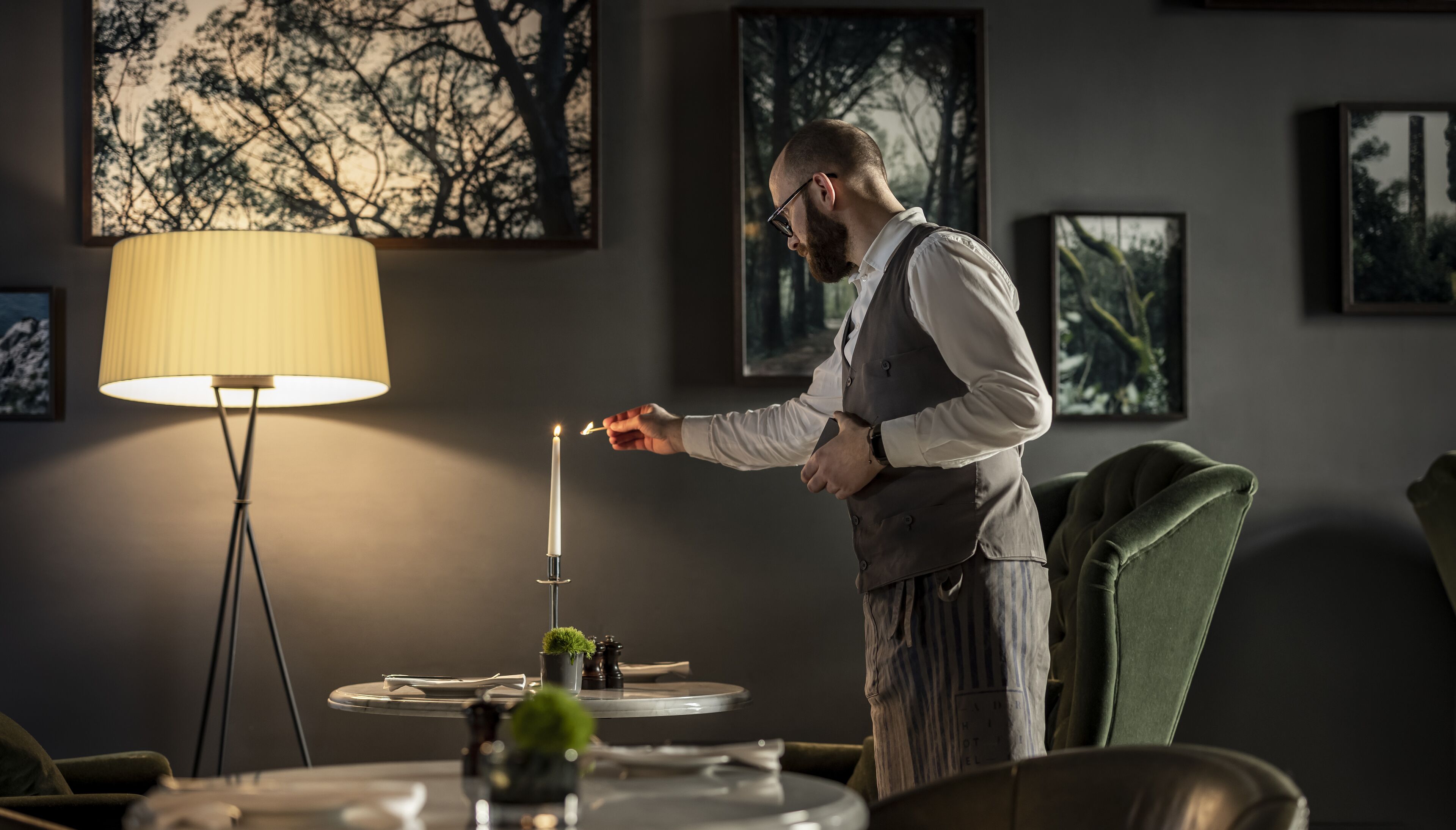 A man lighting a candle in a warmly lit room with framed pictures on the walls and a floor lamp.