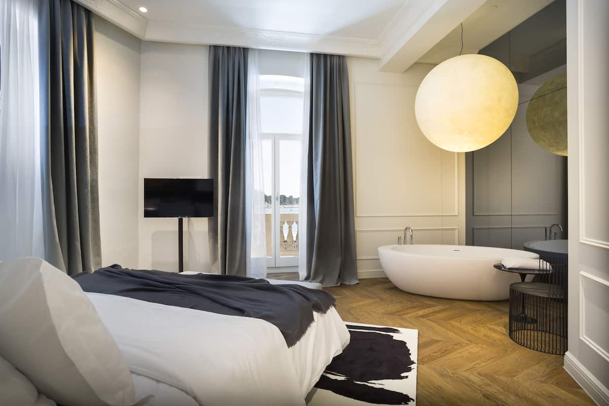 A modern bedroom with a large bed, a flat-screen TV, a bathtub, and a large pendant light.