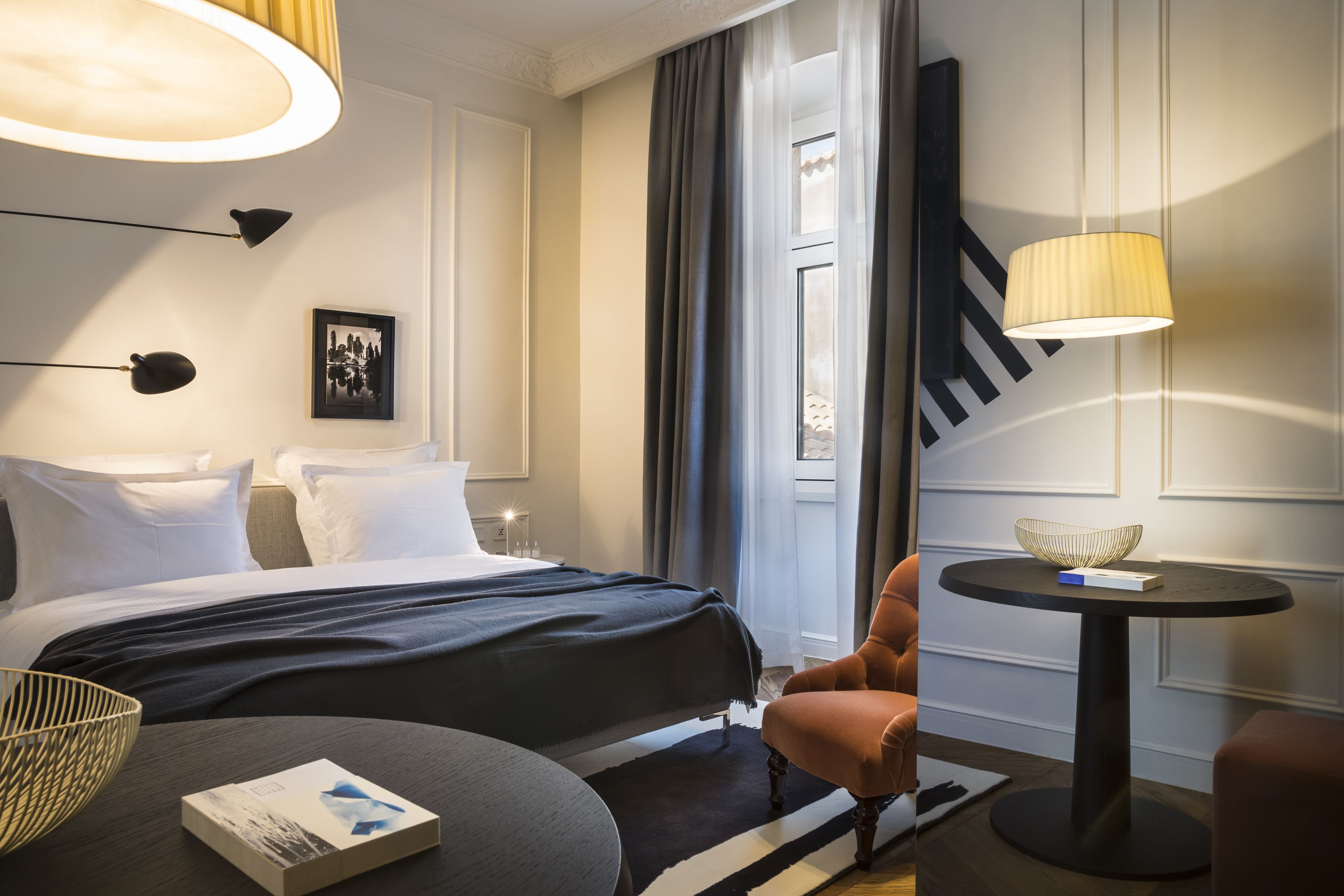 A hotel room with a bed, a round table with a lamp, a chair, and a picture on the wall.