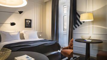 A hotel room with a bed, a round table with a lamp, a chair, and a picture on the wall.