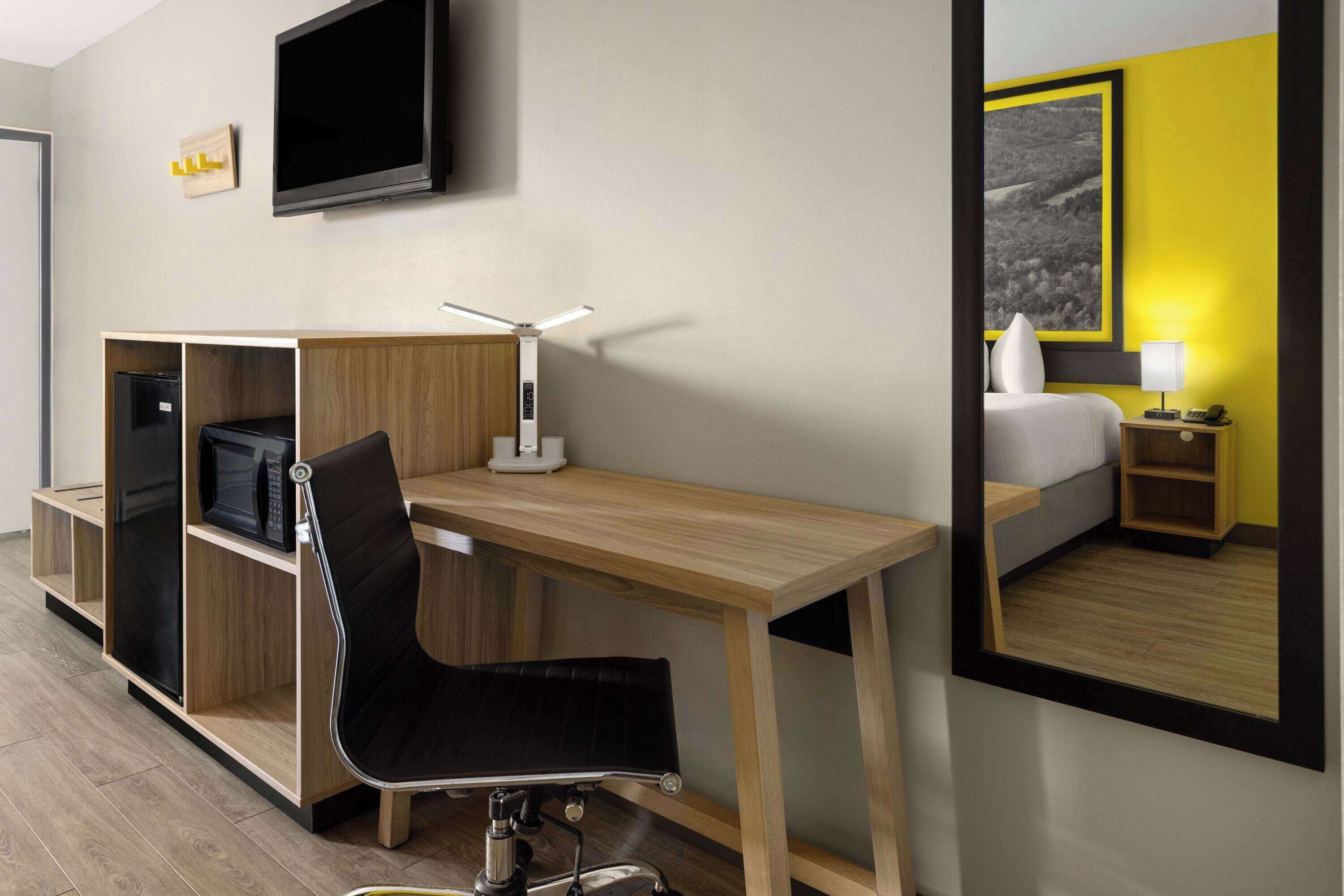 Habitación de hotel con un escritorio de madera, una silla negra, un televisor de pantalla plana, una mesita de noche con lámpara y una cama con una pared de acento amarilla.