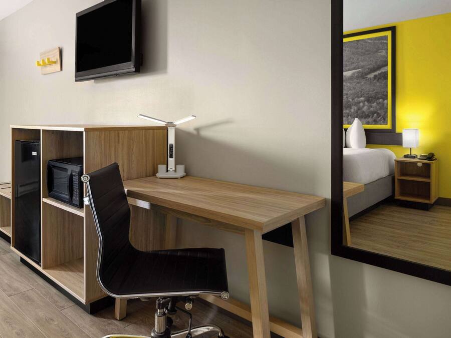 A hotel room with a wooden desk, a black chair, a flat-screen TV, a bedside table with a lamp, and a bed with a yellow accent wall.