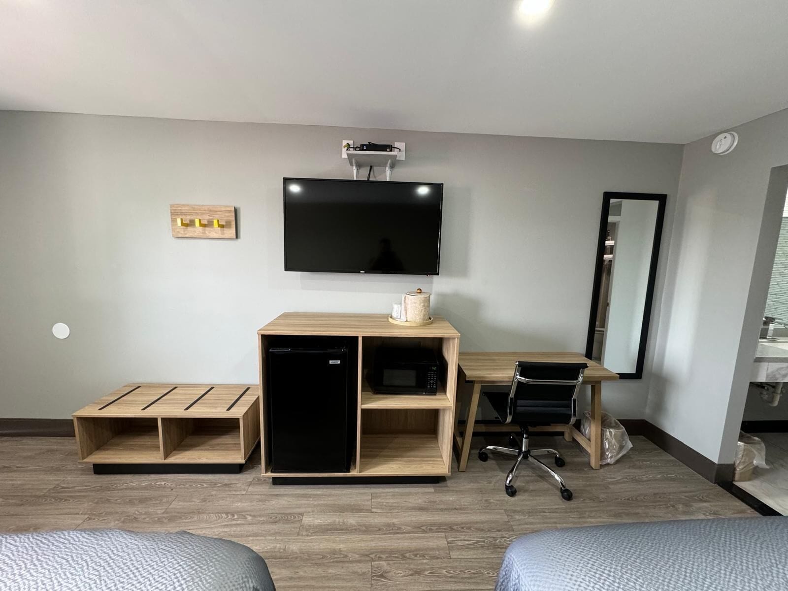 Ein kleines Hotelzimmer mit Flachbildfernseher, Schreibtisch aus Holz, Stuhl, kleinem Kühlschrank und Bett.