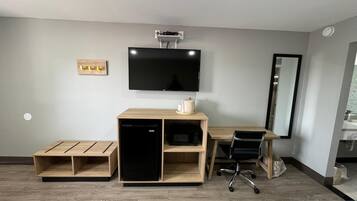 Ein kleines Hotelzimmer mit Flachbildfernseher, Schreibtisch aus Holz, Stuhl, kleinem Kühlschrank und Bett.