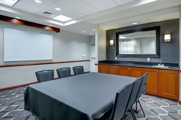 A conference room with a large table, chairs, a projector screen, a kitchen area, and a large mirror.