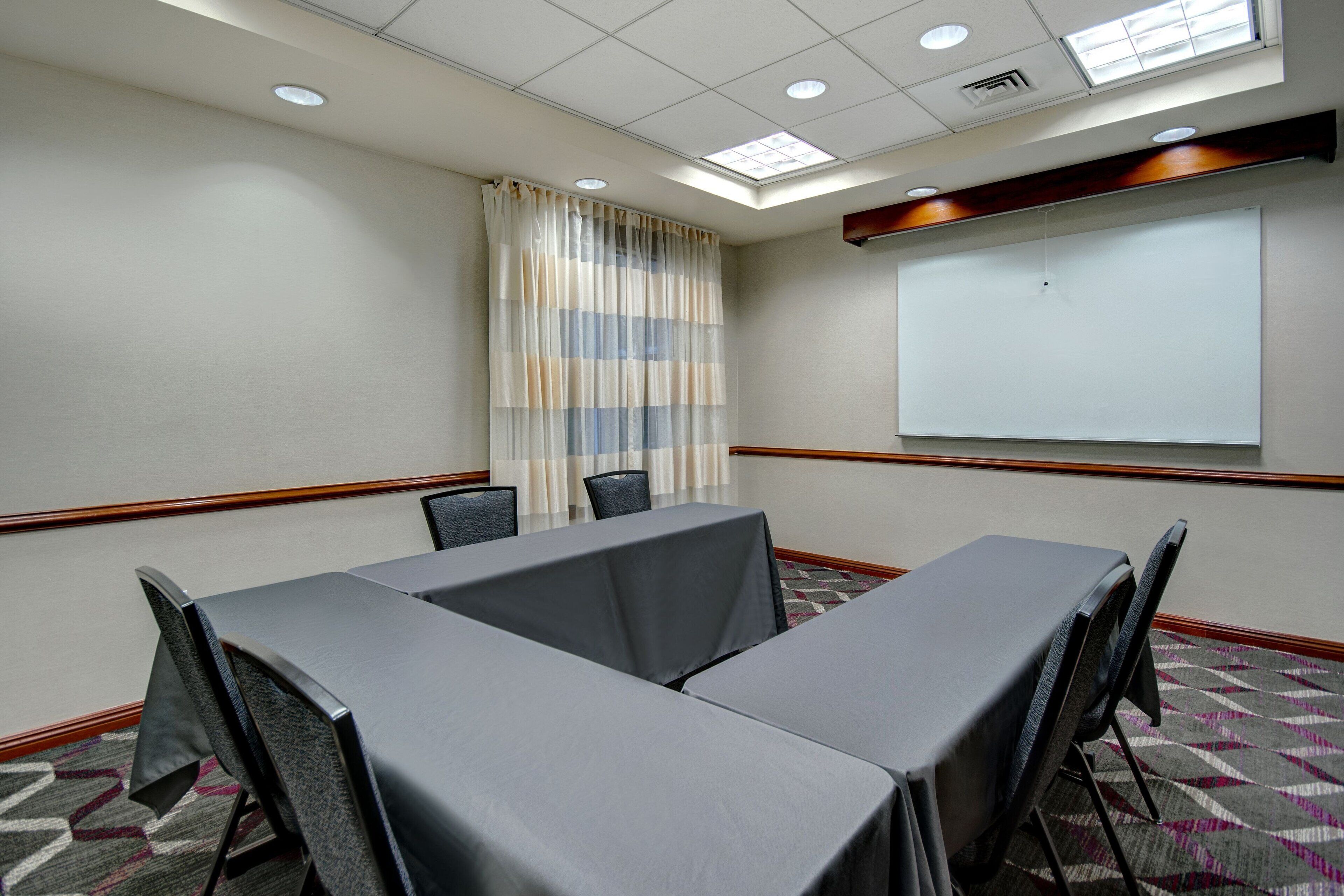 Une salle de conférence équipée d’une grande table, de chaises, d’un écran de projection et d’une fenêtre avec des rideaux.