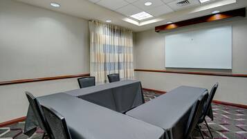 A conference room with a large table, chairs, a projector screen, and a window with curtains.