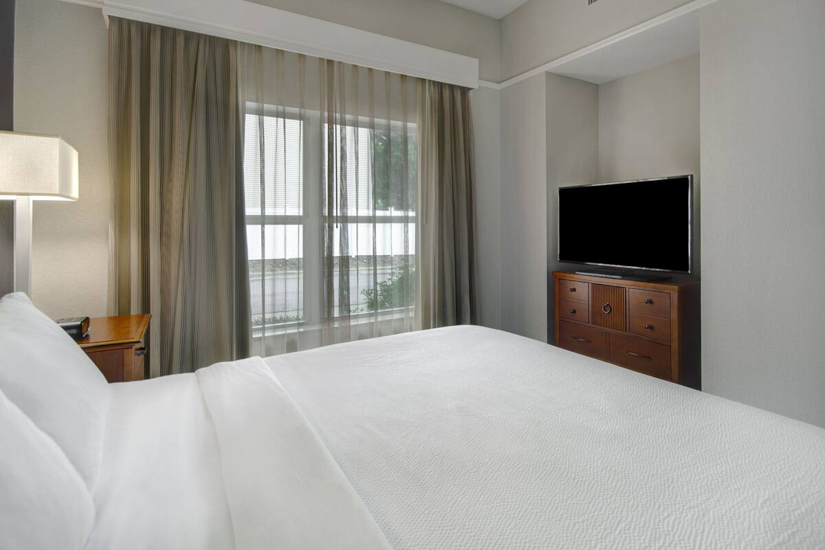 A hotel room with a large bed, a television on a wooden dresser, a lamp, and a window with curtains.
