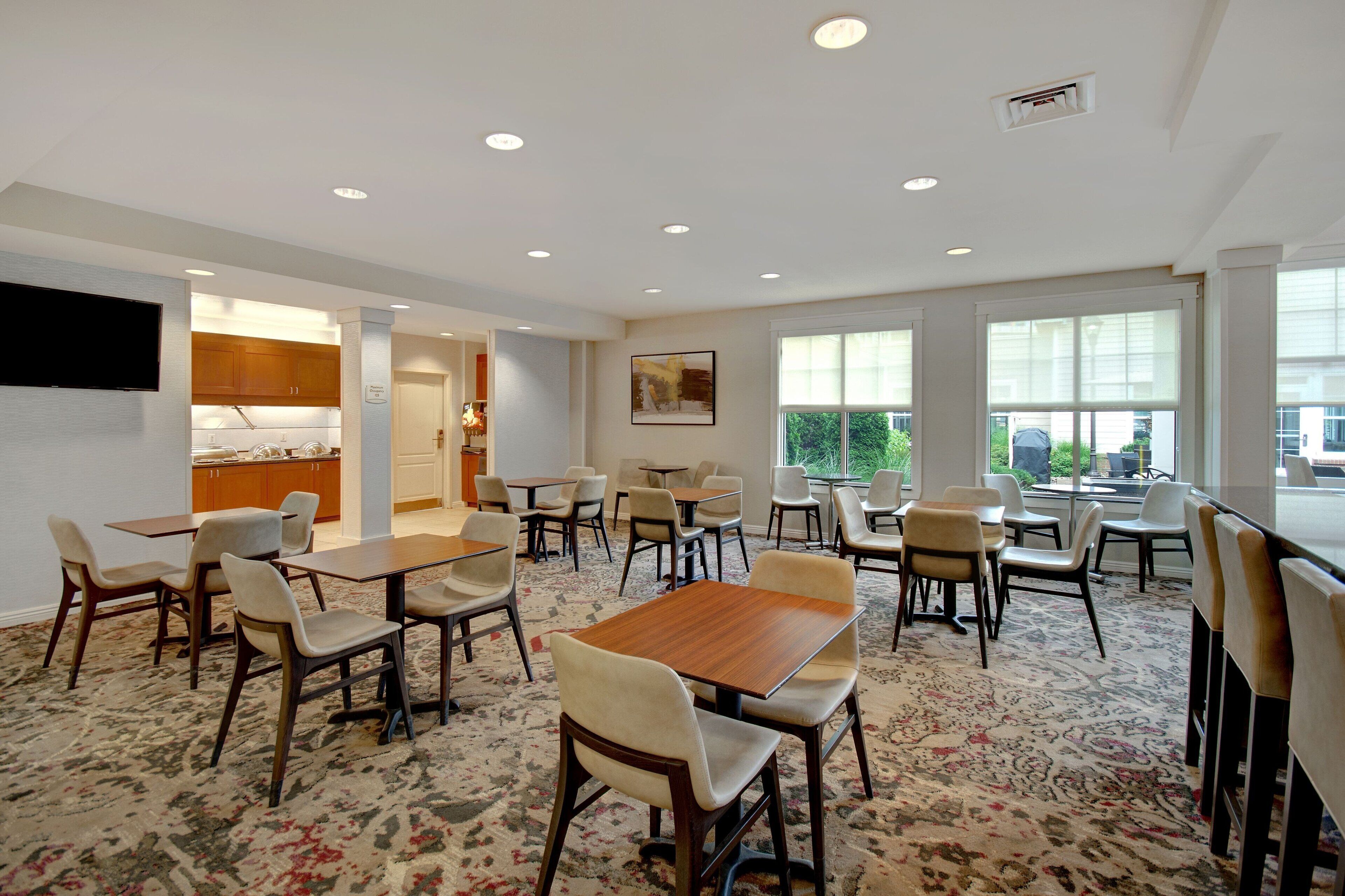 Un grand espace dédié à la salle à manger, avec plusieurs tables et chaises, une cuisine à l’arrière-plan et de grandes fenêtres qui laissent entrer la lumière naturelle.