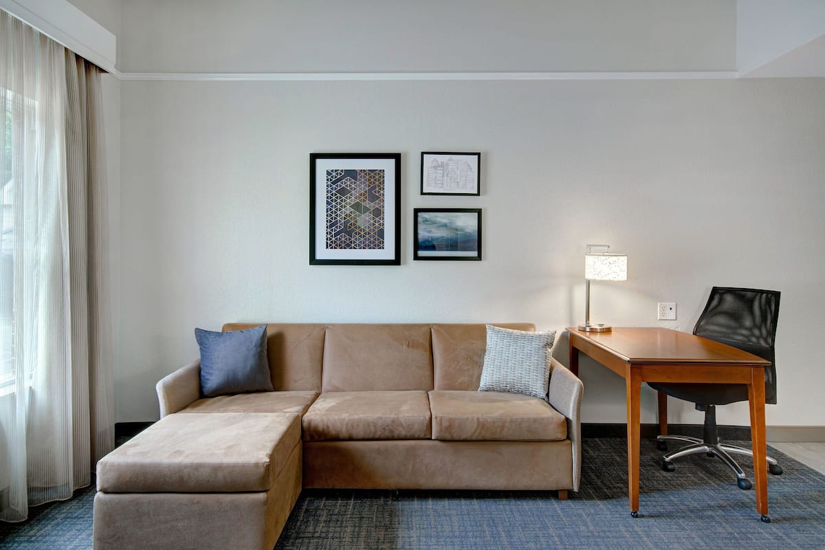 A hotel room with a beige sectional sofa, a wooden desk, and two framed artworks on the wall.