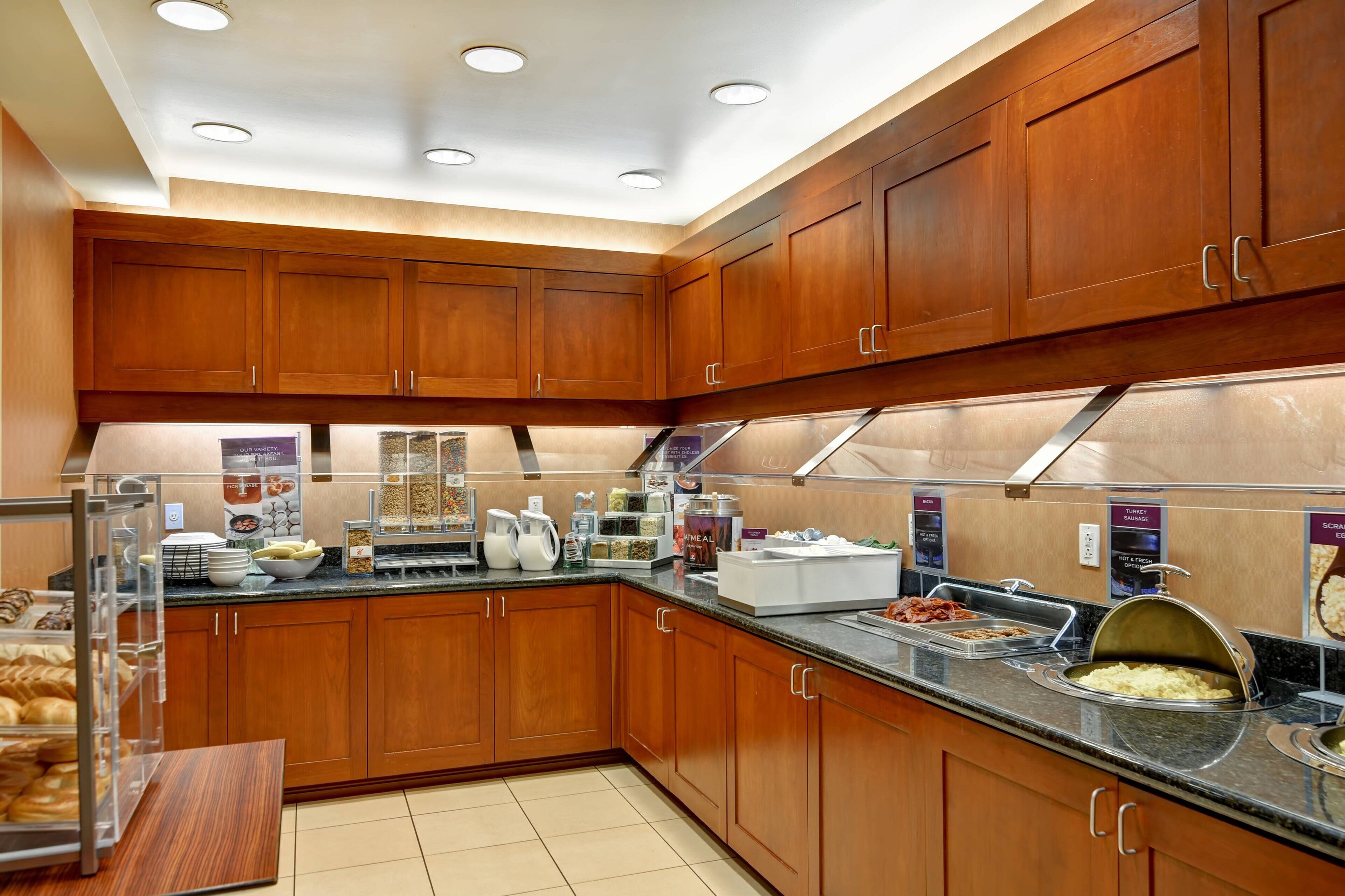 A modern kitchen with wooden cabinets, a granite countertop, and various food items displayed.