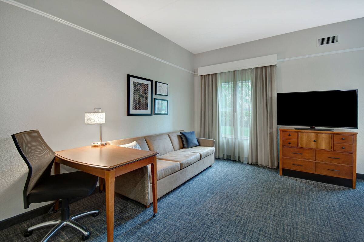 A hotel room with a sofa, a desk, a chair, a television, and a dresser.
