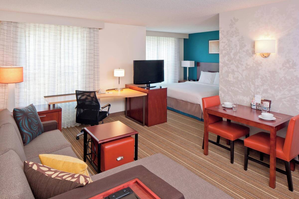 A hotel room with a bed, a desk with a TV, a sofa, and a dining area.