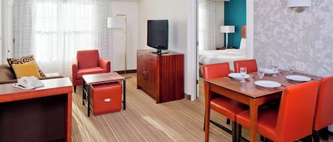A hotel room with a living area, a dining table with chairs, a TV on a wooden cabinet, a bed with a blue wall, and a bathroom visible through an open door.