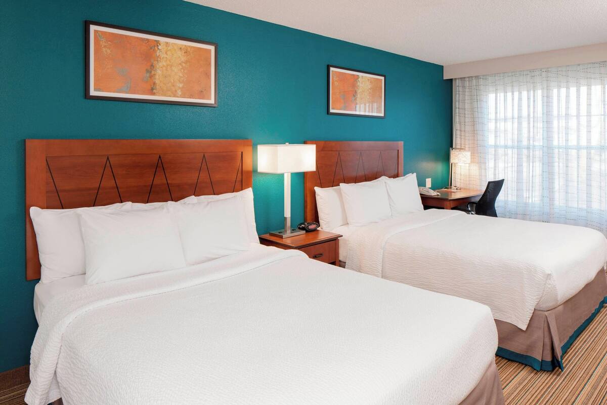 A hotel room with two beds, a nightstand with a lamp, a desk with a chair, and artwork on the walls.