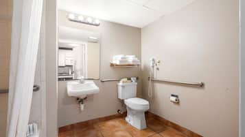 Un baño con lavamanos, inodoro y espejo. Hay barras de apoyo y una cortina de ducha.