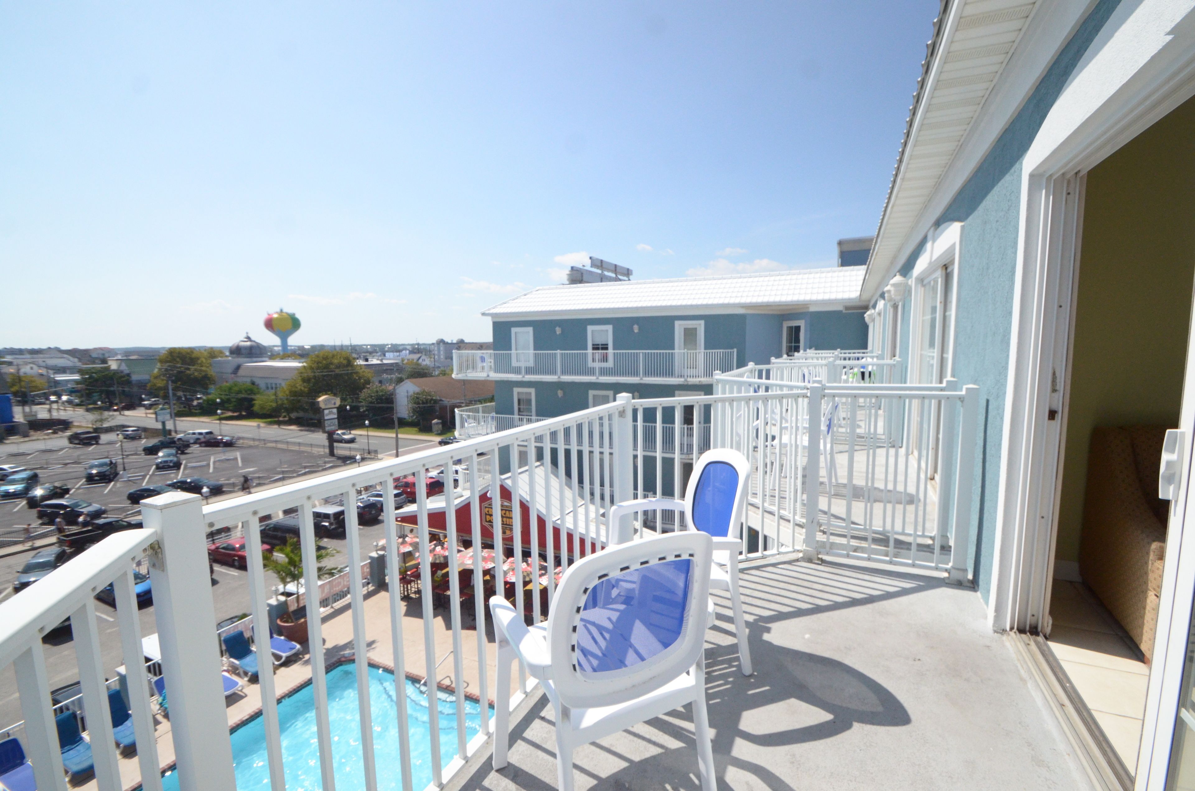 A balcony with a pool, white chairs, and a view of a parking lot and buildings.