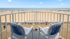 Un balcon avec des balustrades blanches et des chaises bleues, donnant sur une plage où l’on aperçoit des gens et des parasols.