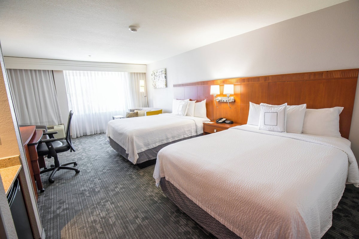 Une chambre d’hôtel avec deux lits, un bureau, une chaise et une fenêtre avec des rideaux.