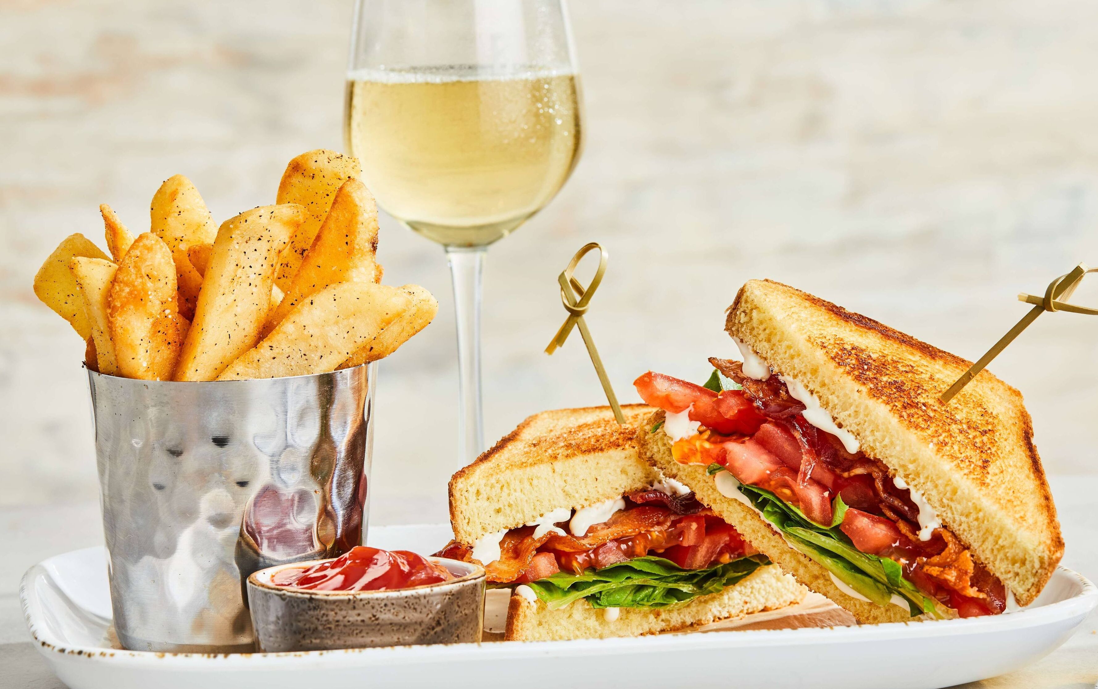 Un sándwich con tocino, tomate y lechuga, una copa de vino blanco y un vaso de papas fritas sazonadas.