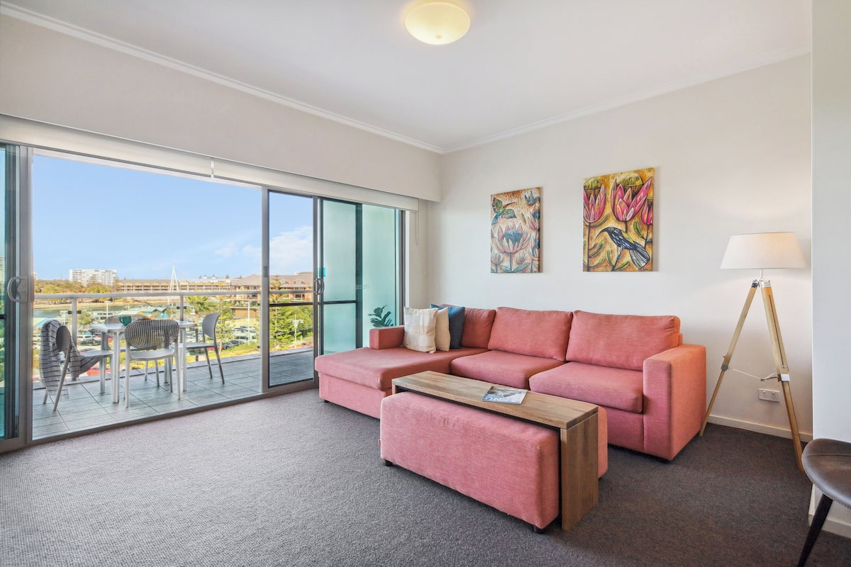 A modern living room with a pink sofa, a wooden coffee table, and a view of the city through large glass doors.