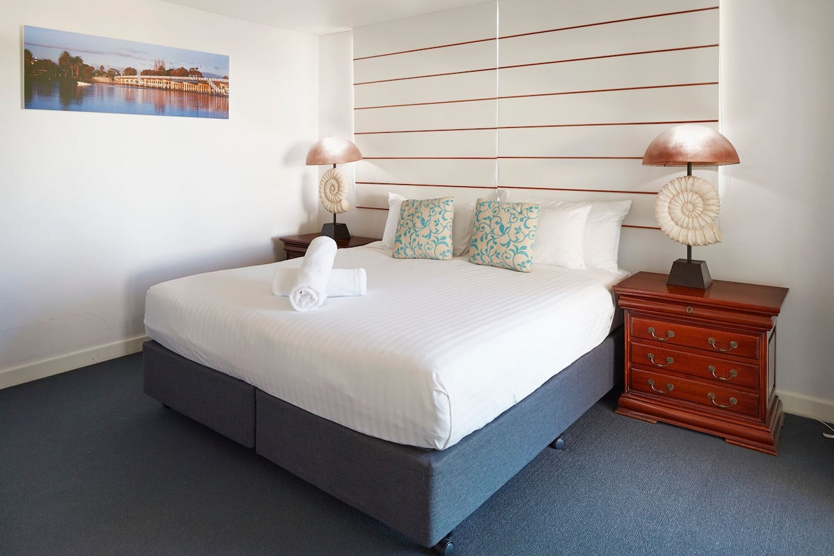A hotel room with a large bed, two bedside lamps, a wooden nightstand, and a wall-mounted headboard.