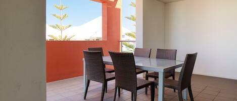 Un comedor con mesa de vidrio y sillas de mimbre, pared roja y vista a palmeras.