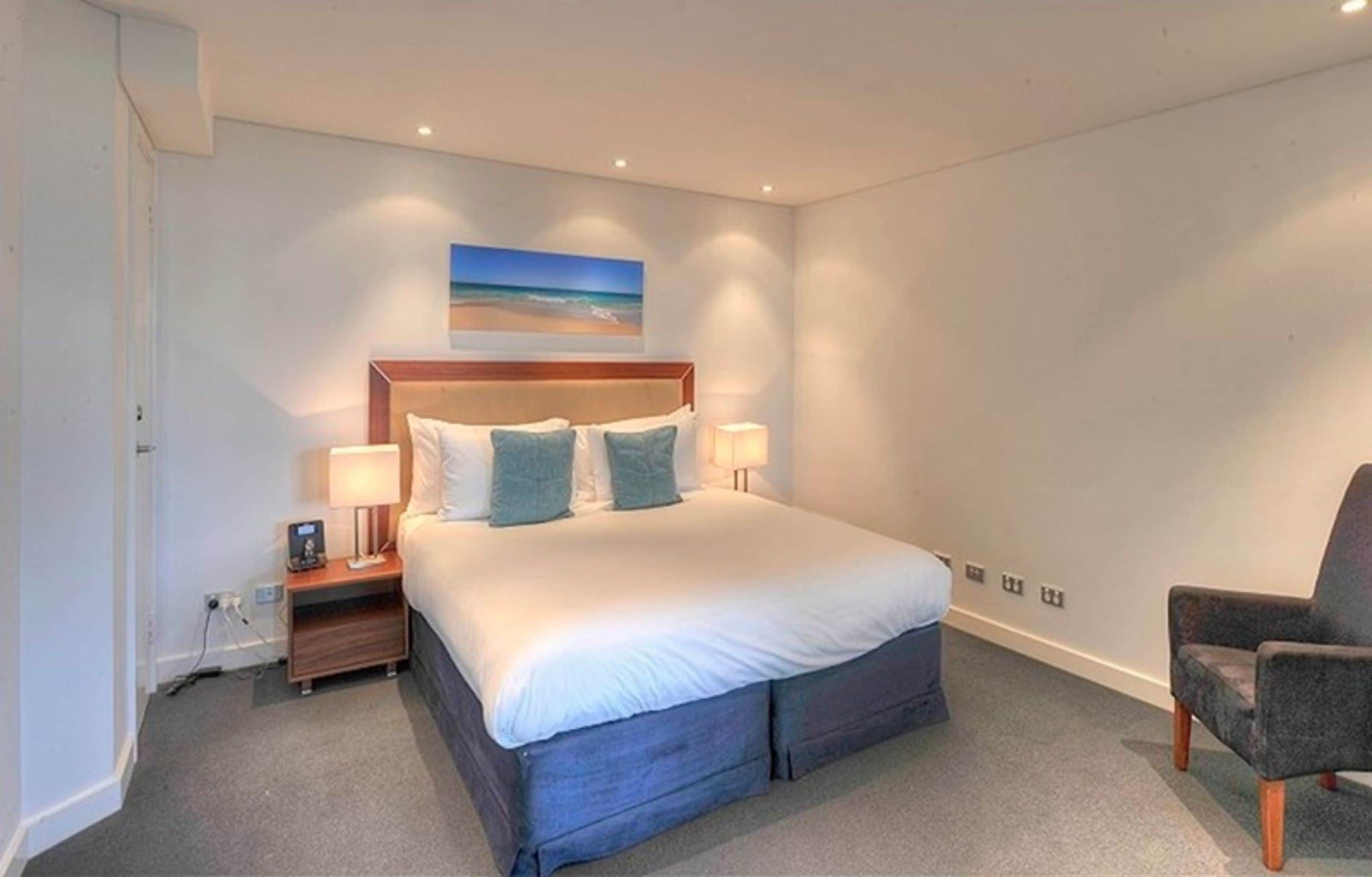 A bedroom with a large bed, bedside lamps, a nightstand, a chair, and a painting on the wall.