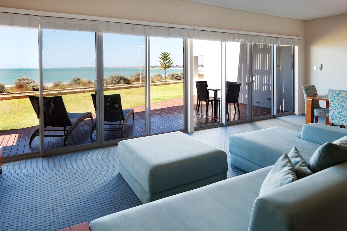 A modern living room with a large sliding glass door leading to a deck with a view of the ocean. There are two sofas and a small table with chairs.