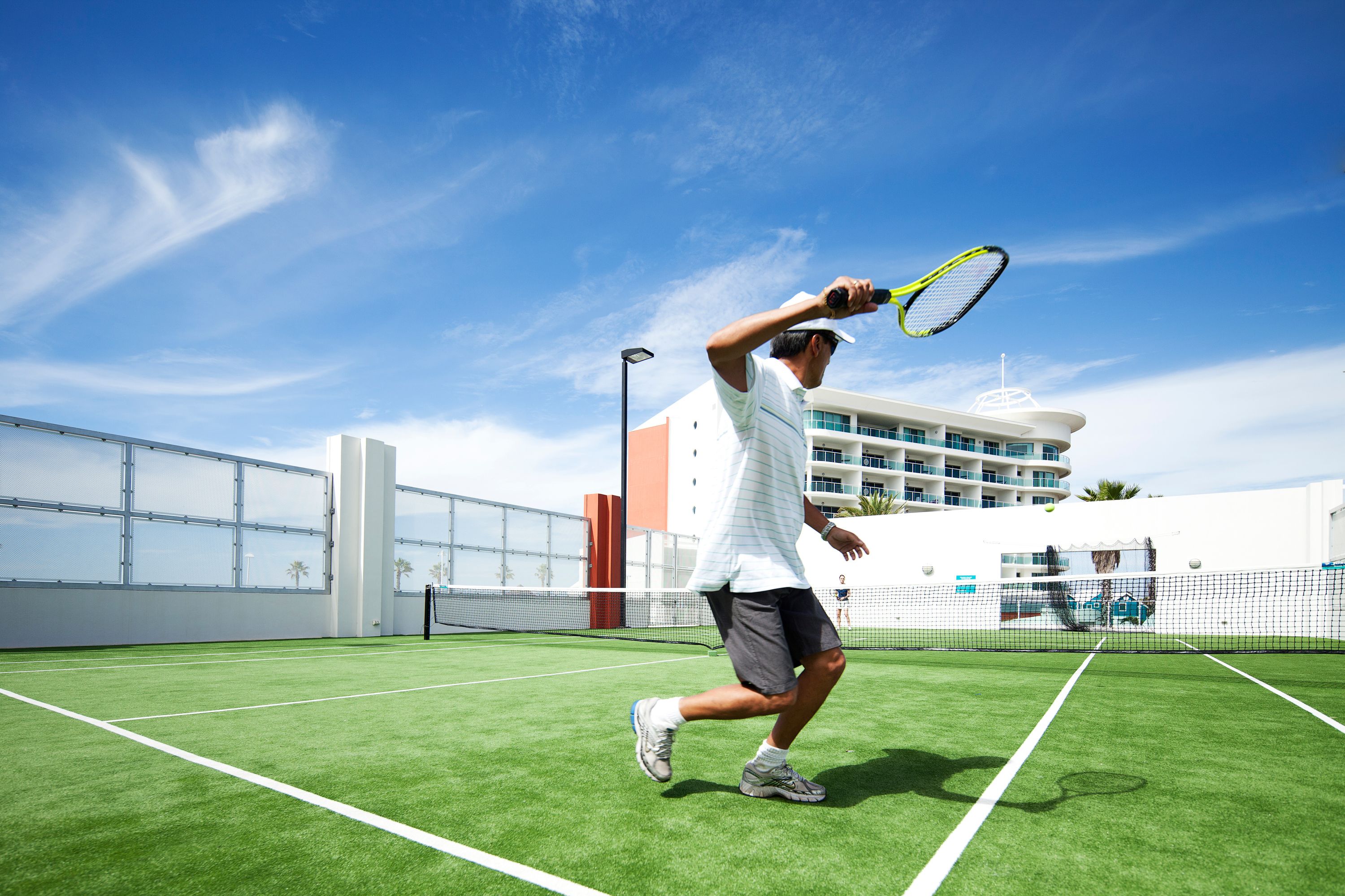 A tennis court with a net, a player in a white shirt and shorts, and a building in the background.