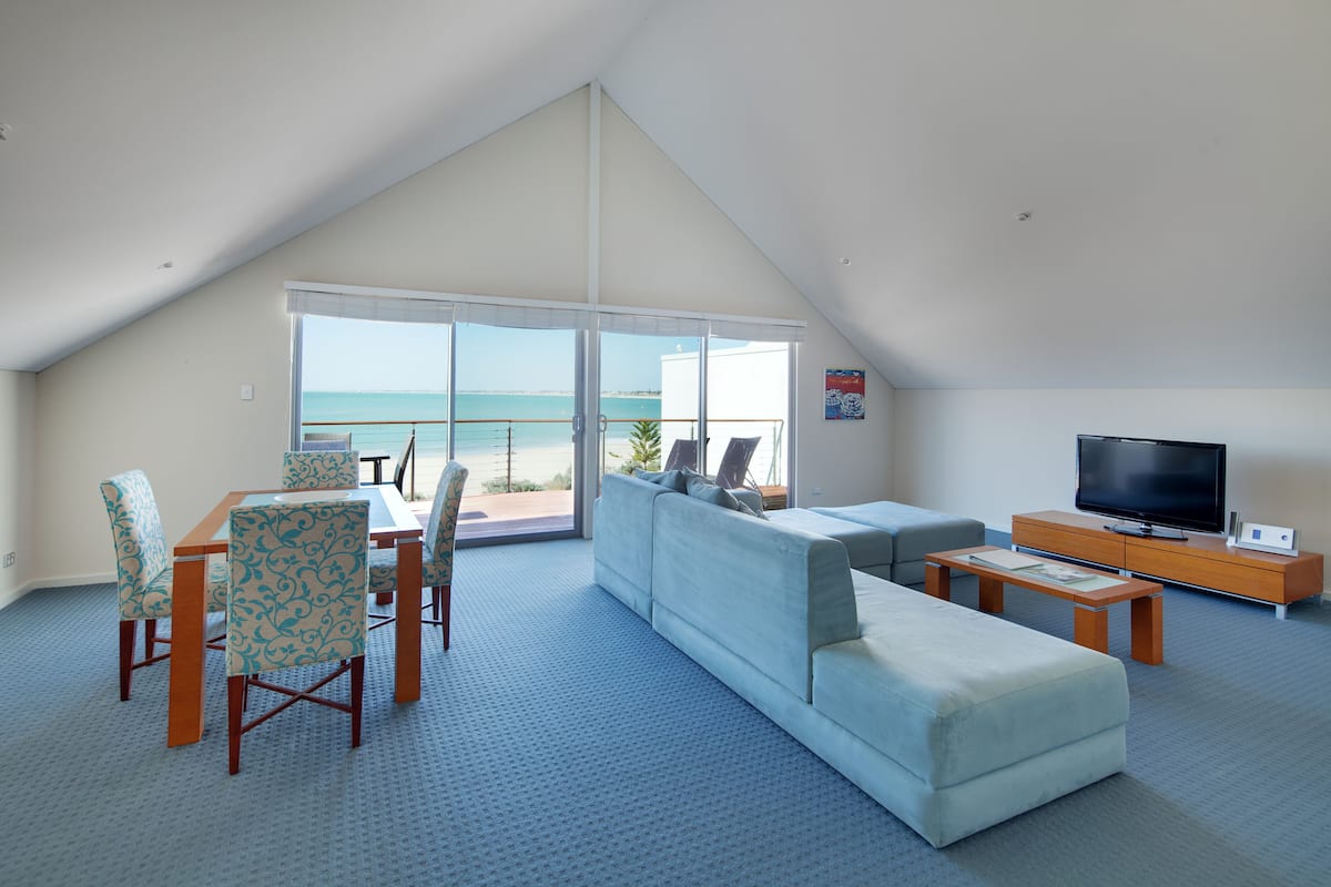 A spacious living area with a large window overlooking the sea, a sofa, a TV, and a dining table with chairs.