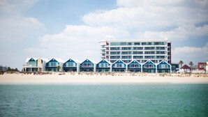 Un hotel frente a la playa con varios edificios de techos azules, un edificio grande y blanco, y una playa de arena.