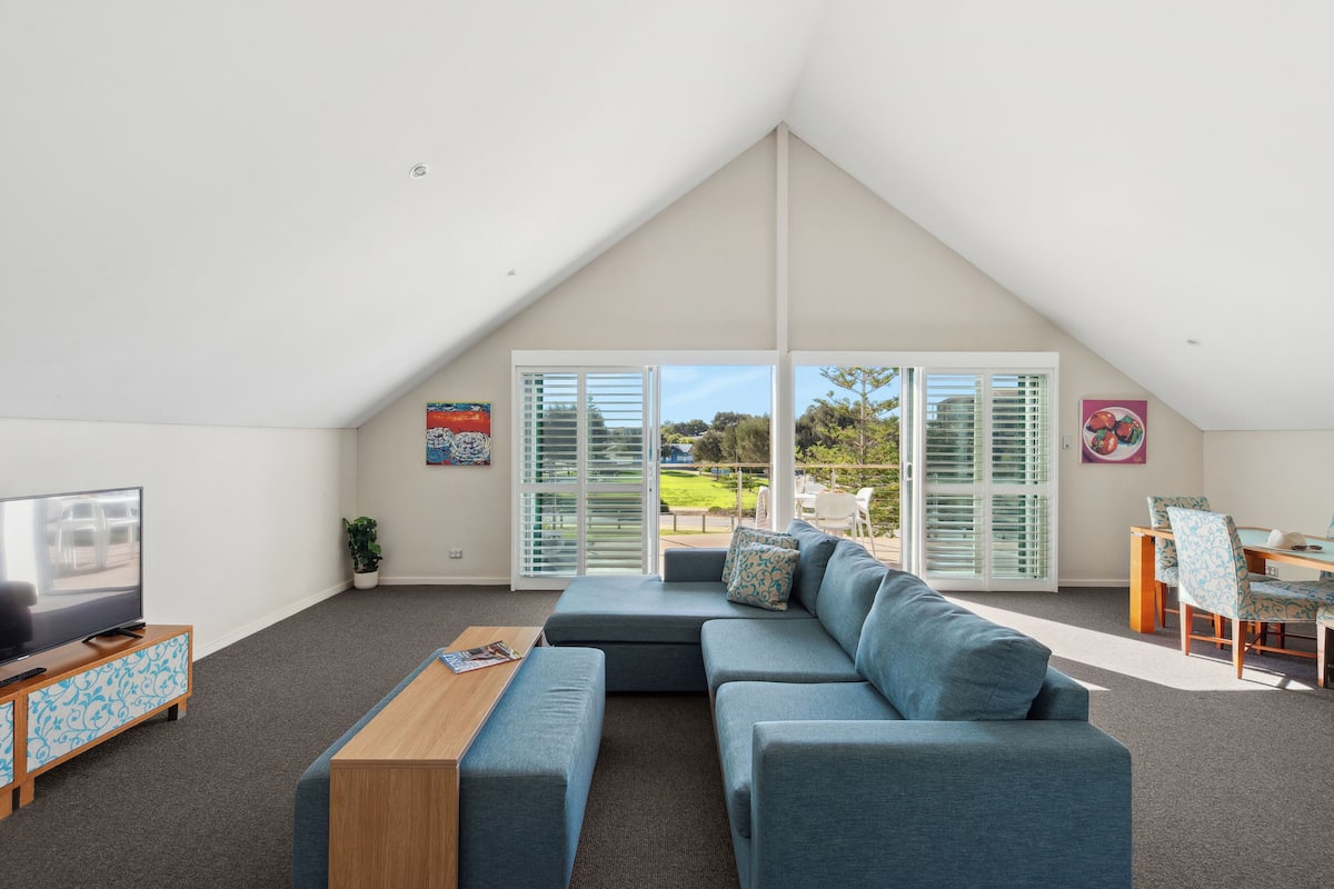 A modern living room with a large sectional sofa, a coffee table, and a TV on a wooden cabinet. There is a dining area with a table and chairs, and a large sliding glass door leading to an outdoor patio.