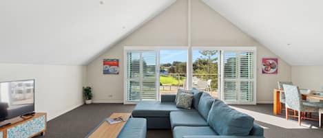 A modern living room with a large sectional sofa, a coffee table, and a TV on a wooden cabinet. There is a dining area with a table and chairs, and a large sliding glass door leading to an outdoor patio.