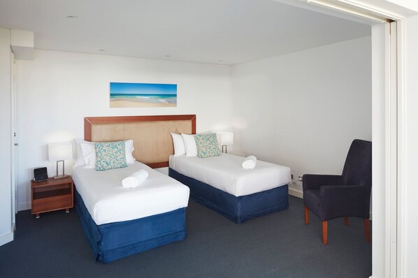 A hotel room with two beds, a desk, a chair, and a painting of a beach.