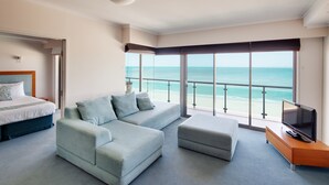 Habitación de hotel con una cama grande, un sofá azul claro, un televisor sobre un soporte de madera y un balcón con vista al océano.