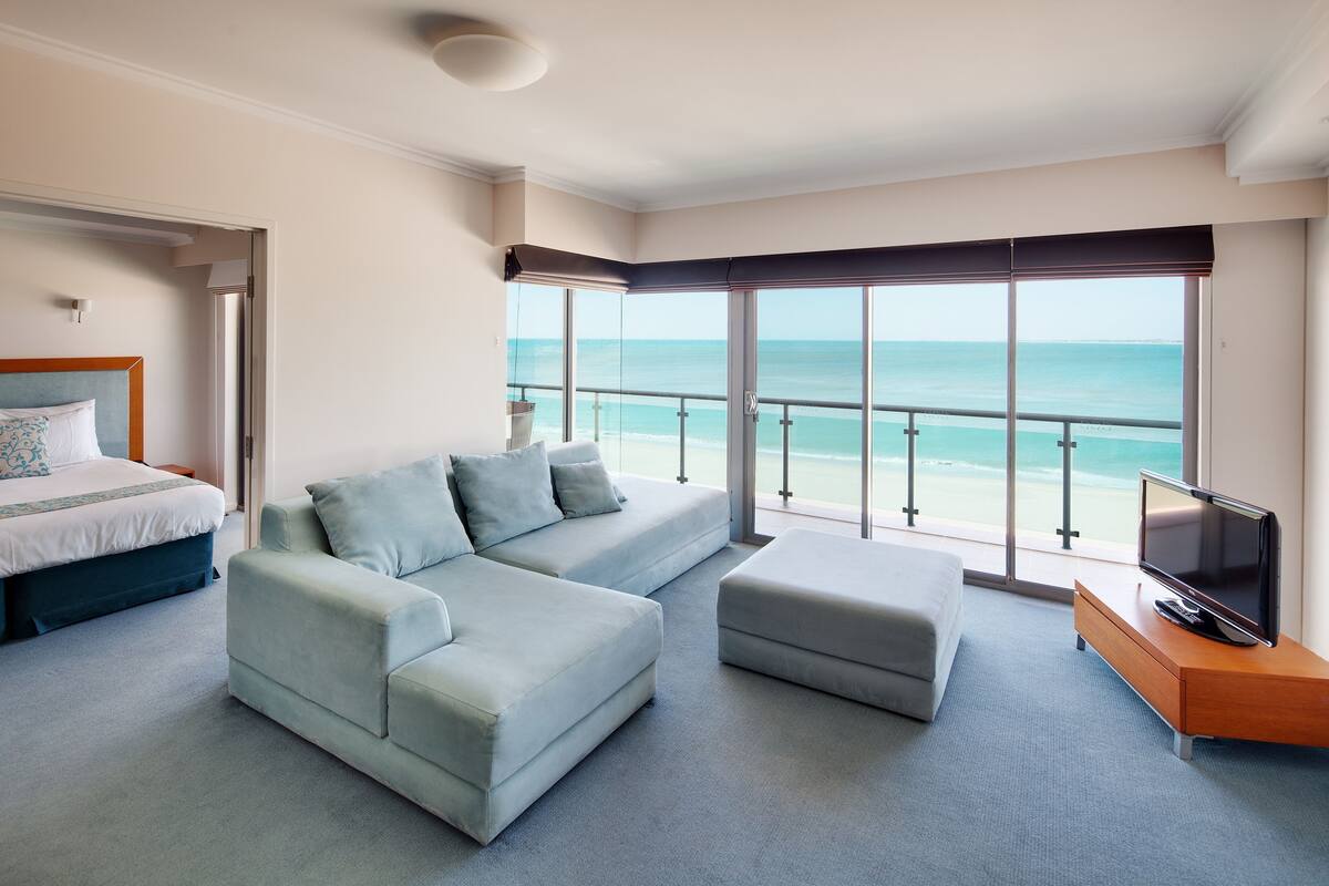 A hotel room with a large bed, a light blue sofa, a TV on a wooden stand, and a balcony with a view of the ocean.
